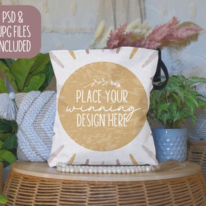May include: A white tote bag with a brown circle and the text "Place your winning design here". The bag is sitting on a wicker table with a white beaded necklace and a white throw pillow.