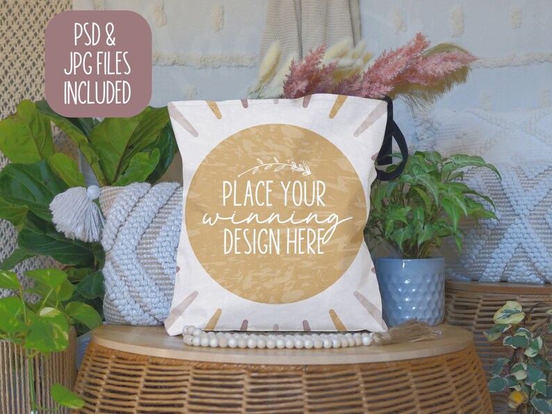 May include: A white tote bag with a brown circle and the text "Place your winning design here". The bag is sitting on a wicker table with a white beaded necklace and a white throw pillow.