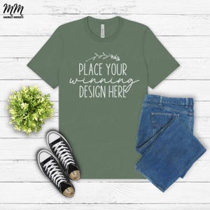 May include: A flat lay mockup of a green t-shirt with the text "Place your winning design here" on it. The t-shirt is on a white wooden surface with a pair of black sneakers, a small green plant, and a pair of blue jeans.