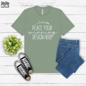 May include: A sage green t-shirt with short sleeves and a crew neck. The shirt has the text "Place your winning design here" printed on the front. The shirt is laid flat on a white wooden surface with a pair of black sneakers and blue jeans.