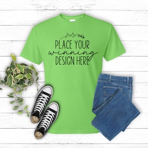 May include: A bright green t-shirt with the text "PLACE YOUR winning DESIGN HERE" in black. The shirt is lying flat on a white wood background with a pair of black sneakers and blue jeans.