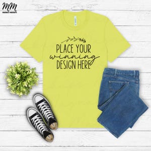 Può includere: Una maglietta gialla a maniche corte e collo rotondo. La maglietta ha la scritta "Place your winning design here" stampata sul davanti. La maglietta è stesa su una superficie di legno bianco con un paio di scarpe da ginnastica nere e un paio di jeans blu.