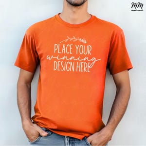 Puede incluir: Una camiseta naranja brillante de manga corta y cuello redondo. La camiseta tiene el texto "Place your winning design here" impreso en blanco en la parte delantera.