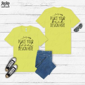 Può includere: Un mockup di una t-shirt gialla con il testo "Place your front design here" sul davanti e "Place your back design here" sul retro. La t-shirt è appoggiata su una superficie di legno bianco con un paio di jeans blu e un paio di scarpe da ginnastica bianche.