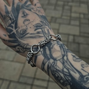 Könnte beinhalten: Ein silberfarbenes Kettenarmband mit zwei großen kreisförmigen Gliedern. Das Armband wird an einem tätowierten Arm getragen. Der Hintergrund ist eine verschwommene Ansicht einer Ziegeloberfläche.