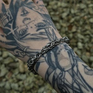 Könnte beinhalten: Ein silberfarbenes Kettenarmband aus kleinen, ineinandergreifenden Metallringen. Das Armband wird an einem Handgelenk mit aufwendigen schwarzen Tätowierungen getragen. Der Hintergrund ist verschwommen, mit kleinen, braunen Kieselsteinen.
