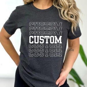 May include: Dark gray t-shirt with the word "CUSTOM" in large white block letters, repeated vertically. The word is outlined in a matching gray color. The shirt is a crew neck style.