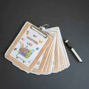 Puede incluir: Un portapapeles marr&oacute;n con una hoja de papel blanca que dice "My Shopping List" y tiene una imagen de un carrito de compras lleno de comestibles. El portapapeles est&aacute; abierto y hay varias hojas de papel en blanco detr&aacute;s de la primera hoja. Un marcador de borrado en seco blanco est&aacute; acostado junto al portapapeles.