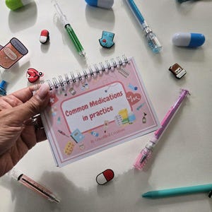 May include: A pink spiral-bound notebook titled "Common Medications in practice" is held by a hand. The image is surrounded by various medical-themed accessories, including pill-shaped charms, syringes, and pens.