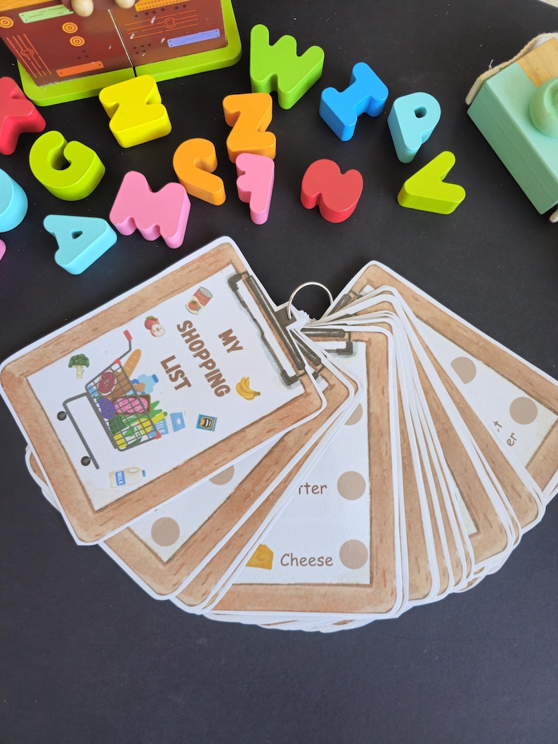 Puede incluir: Un conjunto de bloques de letras de madera de colores y un portapapeles marr&oacute;n con una hoja de papel blanca que dice "Mi lista de la compra" con un carrito de la compra de dibujos animados lleno de comestibles.