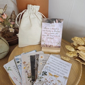 May include: A selection of floral-themed cards with inspirational quotes, presented on a wooden tray. A small drawstring bag and a card holder with a quote are also visible. The cards feature various flower designs and text.