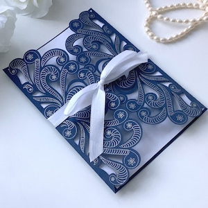 Könnte beinhalten: Lasergeschnittene Hochzeitseinladung in Marineblau mit einem weißen Satinband. Das komplizierte Design zeigt Wirbel und florale Muster.