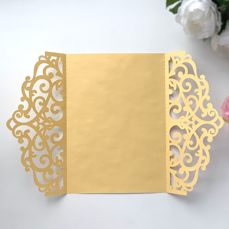 May include: A gold laser cut invitation envelope with a delicate floral design. The envelope is folded in half and the inside is blank.