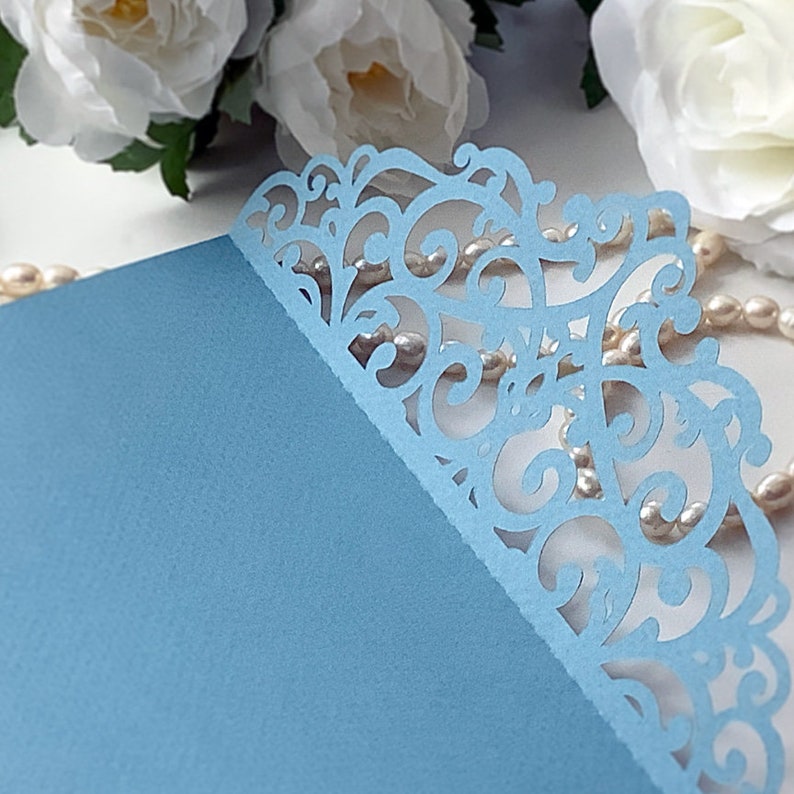 May include: A light blue card with a delicate, intricate lace design cut out of the top right corner. The lace design is a light blue colour and has a floral pattern.