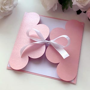 May include: A pink invitation card with a flower-shaped design and a white ribbon tied in a bow. The card has a scalloped edge and a white interior. The overall theme is elegant and suitable for weddings or special events.