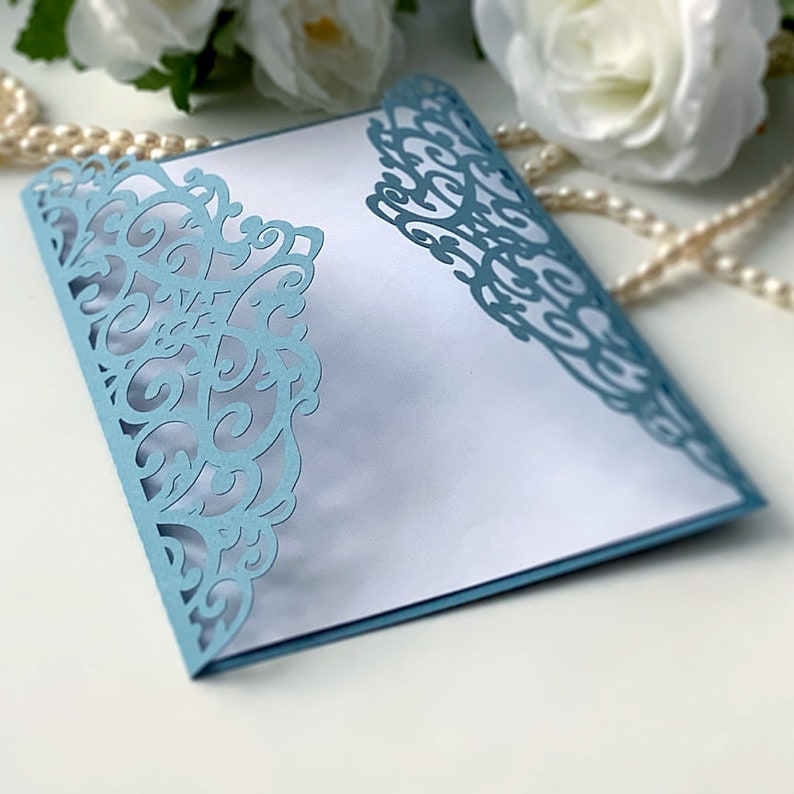 May include: A light blue laser cut envelope with a delicate, intricate floral design. The envelope is folded and has a white interior.