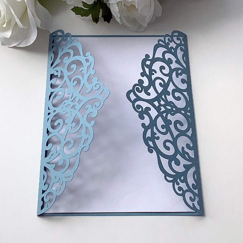 May include: A light blue laser cut invitation envelope featuring a delicate, intricate floral design. The envelope is folded in half and has a white interior.