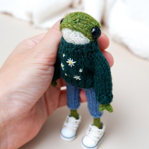 May include: A handmade frog doll wearing a green knit sweater with white daisy accents, blue pants, and white sneakers. The frog has a green head and black eyes. The doll is held in a person's hand.