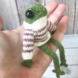 May include: A handmade green frog figurine with a knitted striped sweater in cream and burgundy. The frog has large black eyes and long green legs. The frog is a whimsical, handcrafted item.