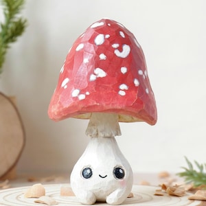 Peut inclure: Une figurine de champignon en bois fantaisiste. Le chapeau rouge est parsemé de taches blanches, et le champignon a un visage souriant, des yeux noirs et un pied blanc. Il est posé sur une tranche de bois, évoquant un thème forestier.
