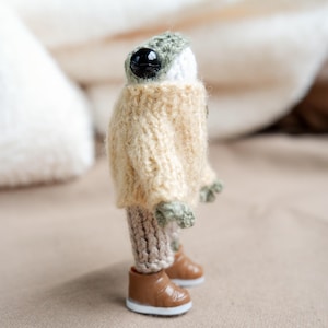 May include: A handmade, miniature creature figurine with a large black eye, wearing a beige sweater, knitted pants, and brown boots. The head is white with a green knitted cap. The figure is standing on a neutral-colored surface.