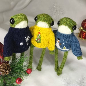 May include: Three handmade frog figurines wearing knitted sweaters. The frogs have green heads, large black eyes, and long green legs. The sweaters are navy blue with a snowflake, yellow with a Christmas tree, and blue with a snowman.