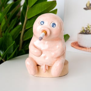 May include: A whimsical, light pink ceramic sculpture of a seated figure with blue eyes and a cigarette in its mouth. The figure is seated on a small, round wooden base. The sculpture has a smooth, glossy finish.