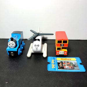 May include: A blue and yellow toy train engine, a white toy helicopter, and a red and yellow double-decker bus toy. The bus has the words "Free the Roads" on the side and "Bulgy" on the front. The toys are on a white surface.