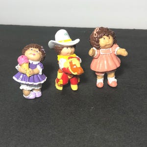 May include: Three Cabbage Patch Kids figurines. One is wearing a purple dress and holding an ice cream cone. Another is wearing a cowboy hat and a red and yellow outfit. The third is wearing a pink dress with a bow in her hair.