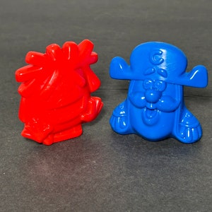May include: Two plastic figurines, one red and one blue. The red figurine is shaped like a person with a large head and a small body. The blue figurine is shaped like a pirate with a hat and a beard.