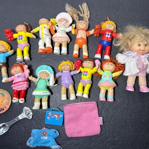 May include: A collection of 13 Cabbage Patch Kids dolls, including a blonde doll with a white coat, a doll with a blue shirt and brown trousers, and a doll with a pink dress. The dolls are all wearing different outfits and have different hairstyles. The dolls are all smiling and looking happy.
