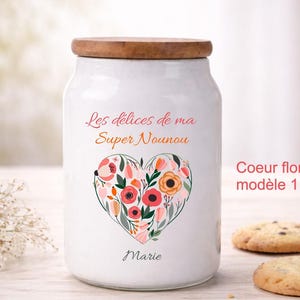 Peut inclure: Un pot en céramique blanc avec un couvercle en bois, orné d'un cœur floral. Le pot porte l'inscription "Les délices de ma Super Nounou" et "Marie". Deux cookies aux pépites de chocolat sont au premier plan.