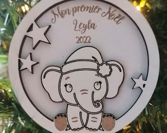 Puede incluir: Adorno navideño de madera blanca con un elefante con gorro de Papá Noel. El adorno tiene el texto "Mon premier Noël Leyla 2022" y recortes de estrellas. Una cuerda está unida para colgarlo.