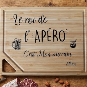 Peut inclure: Planche à découper rectangulaire en bambou avec l'inscription "Le roi de l'APÉRO C'est Mon parrain Ethan" en écriture noire. La planche présente des illustrations d'un mug de bière et d'un verre à cocktail. Olives, salami, amandes et tire-bouchon visibles.