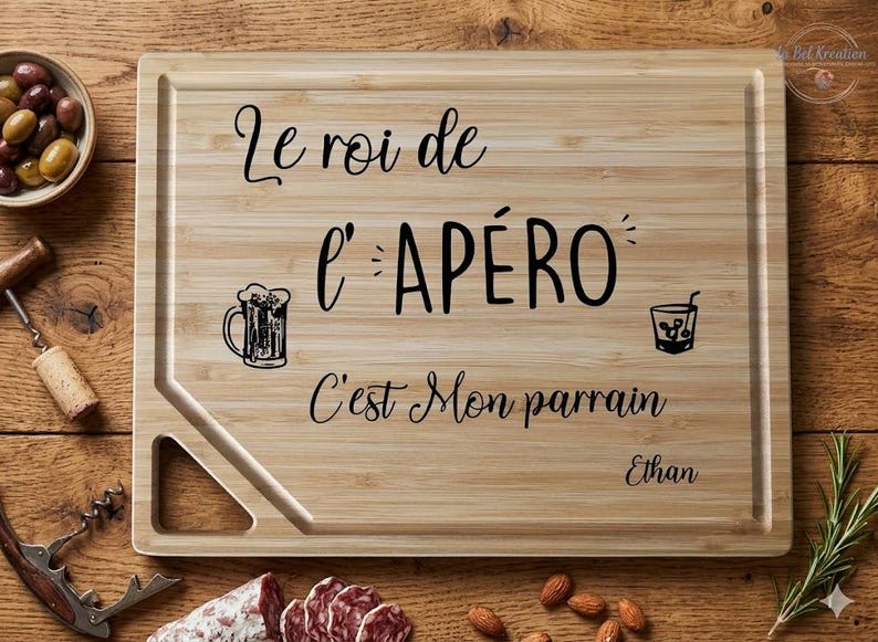 May include: A rectangular bamboo cutting board with the text "Le roi de l'AP&Eacute;RO C'est Mon parrain Ethan" in black script. The board features illustrations of a beer mug and a cocktail glass. Olives, salami, almonds, and a corkscrew are visible.