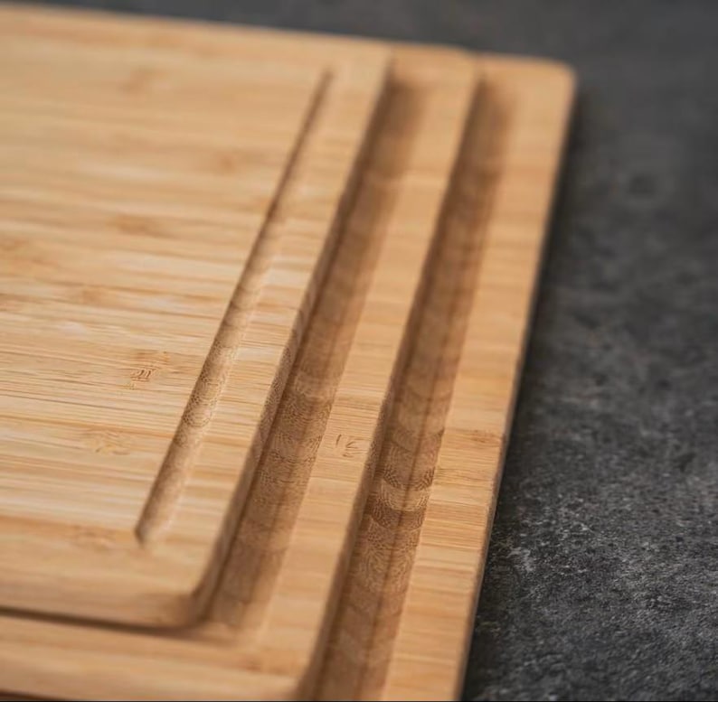 May include: Close-up of a rectangular bamboo cutting board with a juice groove. The board has a natural wood grain pattern and a light brown color. The grooves are carved into the surface of the board.