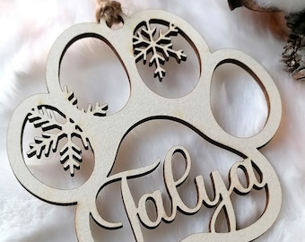 Puede incluir: Un adorno de madera con forma de huella de pata con el nombre "Taliya" recortado y copos de nieve en los lados. El adorno es de color marrón claro y tiene una cuerda de yute para colgar.