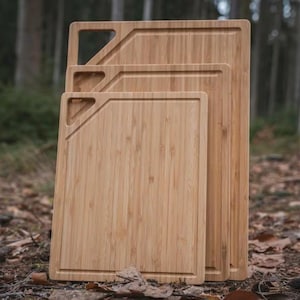 May include: Three bamboo cutting boards of varying sizes, stacked together. Each board has a rectangular shape with a recessed edge and a diagonal cut-out handle. The boards are a light brown color and appear to be made of natural bamboo.