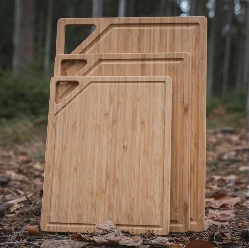 May include: Three bamboo cutting boards of varying sizes, stacked together. Each board has a rectangular shape with a recessed edge and a diagonal cut-out handle. The boards are a light brown color and appear to be made of natural bamboo.