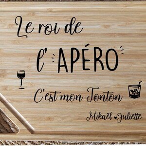 May include: A rectangular bamboo cutting board with a juice groove and a handle. The board features black script that reads "Le roi de l'AP&Eacute;RO C'est mon Tonton Mika&euml;l Juliette" with a wine glass and cocktail illustration.