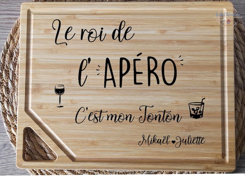 May include: A rectangular bamboo cutting board with a juice groove and a handle. The board features black script that reads "Le roi de l'AP&Eacute;RO C'est mon Tonton Mika&euml;l Juliette" with a wine glass and cocktail illustration.
