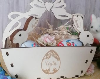 Puede incluir: Una cesta de Pascua de madera blanca con orejas de conejo y un lazo. La cesta está llena de paja, flores y huevos de chocolate. Los nombres "Kymia", "Edward" y "Leyla" están grabados en la cesta.