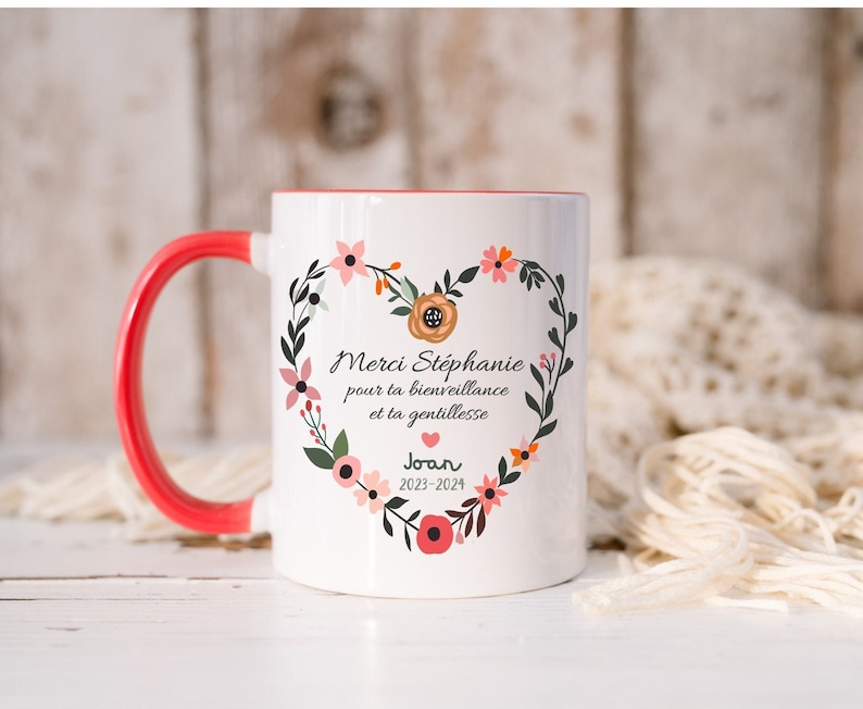 May include: White ceramic mug with a red handle. The mug has a floral design with a heart shape in the center. The text "Merci Stephanie pour ta bienveillance et ta gentillesse Joan 2023-2024" is printed on the mug.