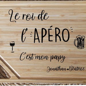 May include: A rectangular wooden cutting board with engraved text and illustrations. The text reads "Le roi de l'AP&Eacute;RO C'est mon papy Jonathan B&eacute;atrice." Includes a wine glass and beer mug illustration.