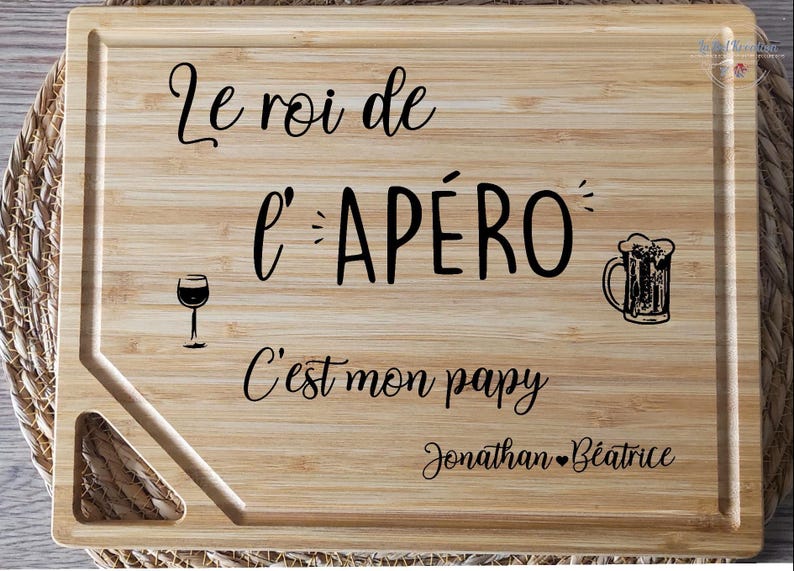 May include: A rectangular wooden cutting board with engraved text and illustrations. The text reads "Le roi de l'AP&Eacute;RO C'est mon papy Jonathan B&eacute;atrice." Includes a wine glass and beer mug illustration.