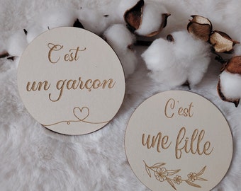 Puede incluir: Dos letreros de madera redondos con el texto "C'est un garçon" y "C'est une fille" sobre un fondo blanco esponjoso. Los letreros son de color madera claro.
