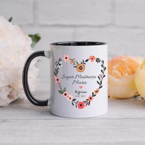 May include: White ceramic mug with a black handle and black rim. The mug has a floral design with a heart in the center. The text on the mug reads "Super Maitresse Marie Alycia 2022-2023".