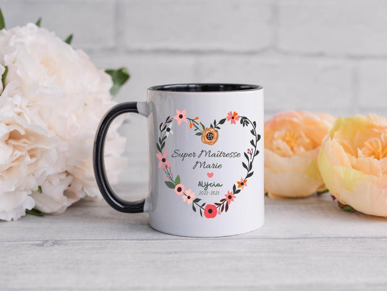 May include: White ceramic mug with a black handle and black rim. The mug has a floral design with a heart in the center. The text on the mug reads "Super Maitresse Marie Alycia 2022-2023".