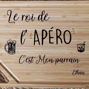 May include: A rectangular wooden cutting board with the text "Le roi de l'AP&Eacute;RO C'est Mon parrain Ethan" in black script. The board features a beer mug and a cocktail glass illustration. The board has a juice groove and a handle.