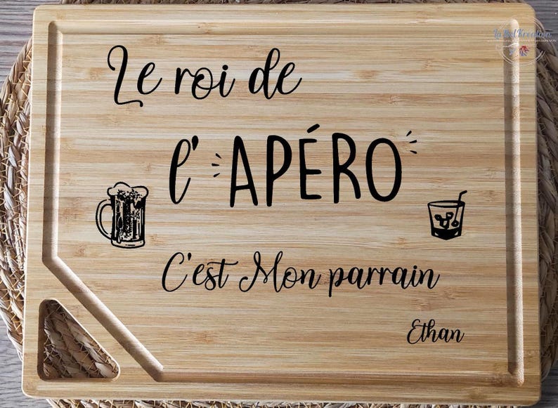 May include: A rectangular wooden cutting board with the text "Le roi de l'AP&Eacute;RO C'est Mon parrain Ethan" in black script. The board features a beer mug and a cocktail glass illustration. The board has a juice groove and a handle.